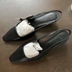Helt nya svarta pumps/kittenheels - Stilrena svarta pumps/kittenheels från H&M med låg klack och spetsig tå. Skorna har öppen häl och smal rem runt hälen. HELT NYA!! Endast testade🔥klack 4cm.