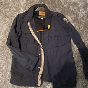 Mörkblå vindjacka från Parajumpers - Säljer en mörkblå vindjacka från Parajumpers med flera fickor och patch på ärmen. Jackan har dragkedja och krage, samt detaljer i beige och orange. Perfekt för blåsiga dagar och har inner ventilation. Nytt skick!! Pris kan diskuteras! 