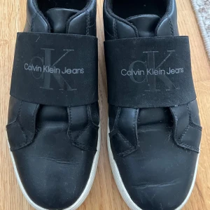 Svarta sneakers från Calvin Klein Jeans - Svarta sneakers från Calvin Klein Jeans med bred resår över foten där loggan syns tydligt. Skorna har vit sula och vit detalj på hälen. Tillverkade i skinnliknande material och har en stilren, sportig look. De har en acceptabelt skick men är sönder vid hälarna. Har beställt de från zalando för 859kr men säljer de för endast 150kr