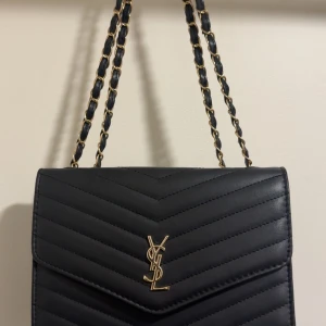 Svart quiltad axelväska från ysl ej äkta - Säljer en svart quiltad axelväska från Yves Saint Laurent ej äkta  med guldfärgade detaljer och kedjerem. Väskan har YSL-logga framtill, magnetlås och flera fack inuti. .
