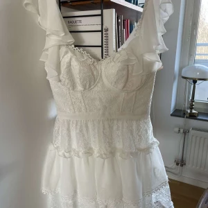 Studentklänning med volang från Koton - Superfin vit klänning från Koton med spetsdetaljer och volanger längs axelbanden. Klänningen har markerad midja, omlottkänsla upptill och flera lager med spets och volang nertill. Perfekt för sommarens tillfällen!