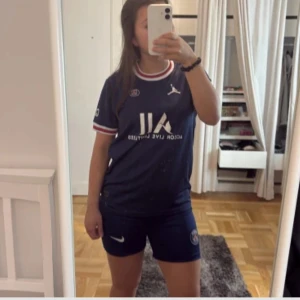 PSG matchtröja dam - Mörkblå PSG fotbollströja med vita och röda detaljer tillsammans med PSG shortsen. Kortärmad modell med klubbmärke och Jordan-logga på bröstet samt sponsortryck framtill. Ribbade muddar i vitt och rött. Perfekt för dig som älskar PSG! Andvönd bara några få gånger max 3 typ. ”Kill” model men funkar för båda tjejer och killar. Man köper t shirten och shortsen tillsammans. 