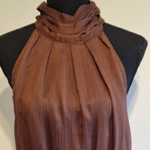 Brun halterneckklänning med rosett från Stockh LM - Säljer en brun halterneckklänning från Stockh LM med hög krage och stor rosett i nacken. Klänningen har ett lätt plisserat tyg och stängs med dragkedja bak. Perfekt för dig som gillar unika detaljer och elegant stil.