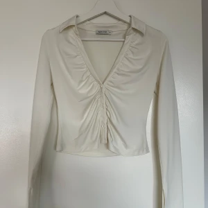 Vit croppad blus från NLY ONE - Säljer en stilren vit croppad blus från NLY ONE med v-ringning och knappar framtill. Blusen har långa ärmar och lätt rynkade detaljer vid bysten. Perfekt för dig som gillar en trendig och feminin look!