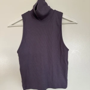Mörklila ribbad polotopp - Säljer en mörklila ärmlös polotopp med ribbad struktur. Toppen har hög krage och är perfekt för att styla lager på lager. Enkel och stilren design som passar till många outfits.