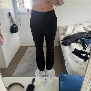 Svarta kostymbyxor med normal passform - Säljer ett par klassiska svarta kostymbyxor med rak passform och hög midja. Byxorna har diskret dragkedja framtill och är tillverkade i ett mjukt material som ger en dressad look. Perfekta för en stilren outfit. Litet igensytt hål i knät men inget som syns