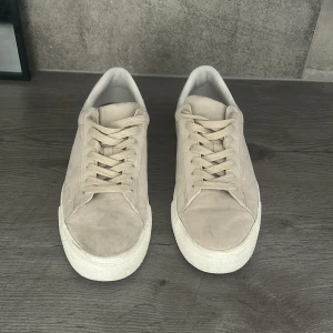 Beige mocka sneakers med vit sula - Säljer ett par stilrena beige sneakers i mocka med klassisk rund tå och platt vit sula. Skorna har snörning och en minimalistisk design som passar till många olika outfits. Perfekta för dig som gillar en clean och enkel look.