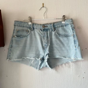 Ljusblå jeansshorts - Säljer ett par ljusblå jeansshorts med råa franskantade benslut och klassisk femficksdesign. Shortsen har hög midja och stängs med knapp och dragkedja. Perfekta nu när sommaren närmar sig 🫶🏻Använd fåtal gånger, storlek M. Midjemått 41 cm💘