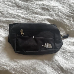 Svart magväska från The North Face - Säljer en svart magväska från The North Face med justerbart band och dragkedja framtill. Perfekt för att bära med sig det viktigaste på ett smidigt sätt. Klassisk logga på framsidan och flera fack för bra organisering.