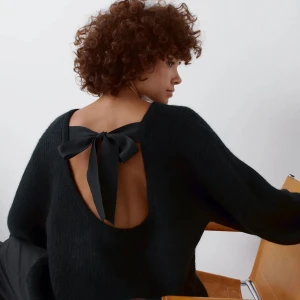 Svart stickad tröja med rosett i ryggen - Säljer en svart stickad tröja med öppen rygg och stor rosett som detalj bak. Tröjan har lång ärm och en avslappnad passform, perfekt för en trendig och mysig look.