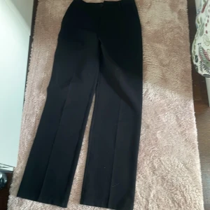 Svarta kostymbyxor från Lindex - Klassiska svarta kostymbyxor från Lindex med raka ben och hög midja. Byxorna har dragkedja och knapp framtill samt hällor för bälte. Perfekta för en stilren och enkel look.