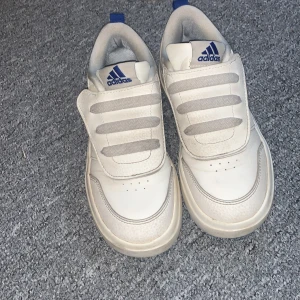 Vita sneakers från Adidas med blå detaljer - Säljer ett par vita Adidas sneakers med blå logga och detaljer. Skorna har kardborreband istället för klassisk snörning och är tillverkade i syntetmaterial. Perfekta för en sportig och avslappnad stil är jätte sköna köpte föra månaden får 600 de är nästan nya ge gärna prisförslag.