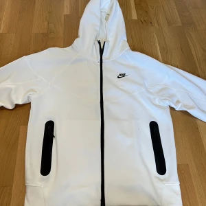 Nike Tech Fleece - Säljer en vit hoodiejacka från Nike med svart dragkedja och svarta detaljer vid fickorna. Jackan har huva och klassisk Nike-logga på bröstet. Perfekt för en sportig och avslappnad stil.