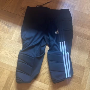 Svarta adidas Tierro fotbollsmålvaktsbyxor - Svarta målvaktsbyxor från Adidas med vadderade partier på knän och höfter. Byxorna har vita ränder längs benen och dragsko i midjan. Perfekta för fotbollsträning eller match.