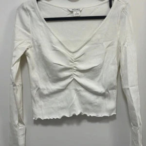 Vit långärmad topp med rynk från Monki - Säljer en vit långärmad topp från Monki med v-ringning och snygg rynkdetalj framtill. Toppen har croppad passform och vågig kant nertill. Perfekt att styla till jeans eller kjol!