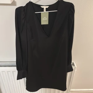 Svart klänning med v-ringning från H&M - Stilren svart klänning från H&M med djup v-ringning och långa ärmar med breda manschetter. Klänningen har lätt puff vid axlarna och är tillverkad i ett slätt polyestertyg. Perfekt för dig som gillar enkel men elegant stil.