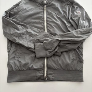 Grå vindjacka från Moncler - Säljer en stilren grå vindjacka från Moncler med klassisk logotyp på ärmen. Jackan har dragkedja framtill, ribbade muddar och krage samt två fickor. Perfekt för dig som gillar enkel och sportig stil.