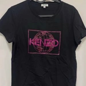 Svart t-shirt från Kenzo med rosa tryck - Svart t-shirt från Kenzo med ett stort rosa tryck framtill som visar varumärkets logga och en glob. Klassisk rund halsringning och korta ärmar. Väldigt liten i storleken, mer som en barn M! Tillverkad i mjuk bomull för en bekväm känsla.