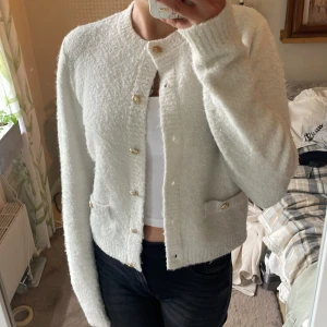 Vit kofta från Holly & Whyte - Säljer en fluffig vit kofta från Holly & Whyte med guldknappar och två små fickor framtill. Koftan har lång ärm och är perfekt att slänga över en topp, har använts ett fåtal gånger. Köpte i vintras för 600kr 