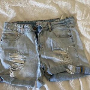 Ljusblå jeansshorts med slitningar från Amisu - Ljusblå jeansshorts med råa slitningar framtill och uppvikta benslut. Klassisk femficksmodell med bälteshällor och knappgylf. Perfekta för en avslappnad stil.