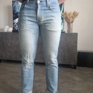 Ljusblå jeansbyxor från Levi's - Säljer ett par klassiska ljusblå jeans från Levi's med normal passform och raka ben. Jeansen har fem fickor, knappgylf och subtila slitningar framtill. Perfekta för en avslappnad stil.