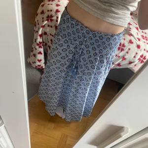 Blåvit långkjol med mönster - Säljer denna långkjol i blått och vitt med dekorativt mönster och smock i midjan. Kjolen har knytband med tofsar framtill. Luftigt och skönt material