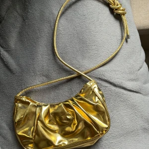Guldig axelväska med metallic-finish - Säljer en snygg axelväska i metallic-guld med rynkad design och smal axelrem. Väskan har magnetlås och ljus insida. Perfekt för att lyfta vilken outfit som helst med lite extra bling.