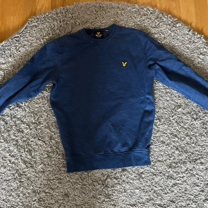 Blå sweatshirt från Lyle & Scott - Säljer en blå sweatshirt från Lyle & Scott med klassisk rund halsringning och gul logga broderad på bröstet. Tröjan har långa ärmar och ribbade muddar vid ärmslut och nederkant. Perfekt för en avslappnad stil.