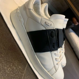 Valentino Open  - Säljer ett par snygga Valentino Open sneakers i vitt skinn med en bred svart rem över mitten. Klassisk och stilren design med vita snören och rund tå. Perfekta för dig som gillar exklusiva och trendiga sneakers.