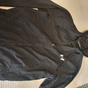 Under Armour jacka - Svart vindjacka från Under Armour med huva och dragkedja. Jackan har en sportig look med logga på bröstet. 