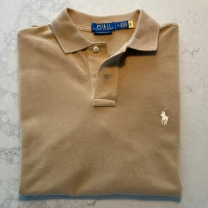 Beige pikétröja från Polo Ralph Lauren - Säljer en beige pikétröja från Polo Ralph Lauren i modellen Custom Slim Fit. Tröjan har klassisk krage, knappar vid halsen och den ikoniska broderade loggan på bröstet. Perfekt för en stilren och avslappnad look.