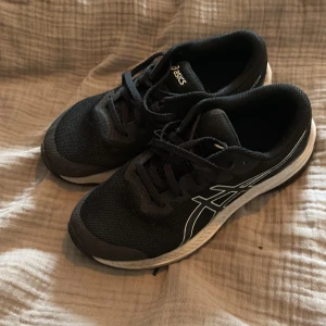 Svarta Asics löparskor - Säljer ett par svarta Asics sneakers med vit sula och vita detaljer på sidan. Skorna har snörning och är tillverkade i syntetmaterial med mesh-överdel för bra andningsförmåga. Sparsamt använda. US 5