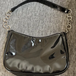 Svart axelväska med kedjedetaljer - Snygg svart axelväska i glansigt syntetmaterial med silvriga kedjedetaljer på axelremmen. Väskan har dragkedja upptill och en stilren, modern look som passar till många outfits.