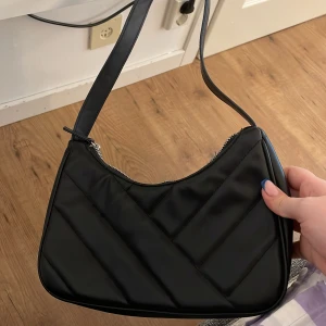 Svart quiltad axelväska - Snygg svart axelväska med quiltad design och dragkedja upptill. Väskan har en stilren form och ett längre axelband som gör den enkel att bära. Perfekt för att lyfta vilken outfit som helst.
