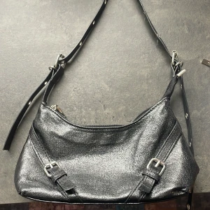 Svart glittrig axelväska med spännen - Snygg svart axelväska med silvriga detaljer. Väskan har justerbart axelband med nitar och två dekorativa spännen framtill. Skriv om det finns några frågor eller för fler bilder!☺️