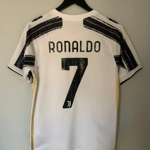 Juventus fotbollströja Ronaldo 7 - Juventus hemmatröja med Ronaldo 7-tryck på ryggen. Tröjan är vit med svarta ränder och guldiga detaljer, Jeep-logga på bröstet och Juventus klubbmärke. Klassisk V-ringning och korta ärmar. Den är ifrån 2020/21 säsongen. 