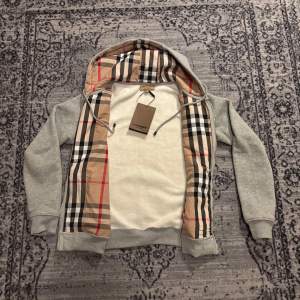 Säljer en grå hoodie med dragkedja från Burberry. Insidan av huvan har det klassiska Burberry-rutiga fodret i beige, svart, rött och vitt. Jackan har broderad logga på bröstet och två fickor framtill.