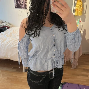 Blå- och vitrandig blus med offshoulder och knappar - Säljer en blå- och vitrandig blus med offshoulder och smala axelband. Blusen har knappar framtill, dragsko i midjan och långa ärmar med öppna axlar. Perfekt för vår och sommar!🩵