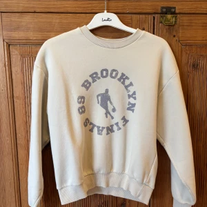 Beige sweatshirt från Gina Tricot med Brooklyn-print - Säljer en beige sweatshirt från Gina Tricot med tryck i grått framtill: 'Brooklyn Finals 89' och en basketspelare. Tröjan har rund halsringning, långa ärmar och ribbade muddar vid ärmslut och nederkant. Storlek xxs.
