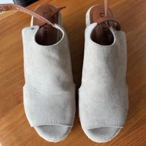 Beige sandaler med kilklack och bruna remmar - Snygga beige sandaler från LINEAR med öppen tå och kilklack i repdesign. Ovandel i mocka och justerbara bruna remmar i syntet med spänne. Perfekta för varma dagar och ger en avslappnad men stilren look.