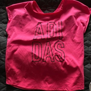 Rosa t-shirt från Adidas - Säljer en rosa t-shirt från Adidas med stor svart logga framtill. T-shirten har korta ärmar och en avslappnad passform. Perfekt för träning eller en sportig vardagslook.