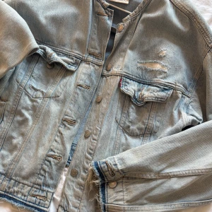 Ljusblå jeansjacka från Levi's - Säljer en klassisk ljusblå jeansjacka från Levi's i storlek M. Jackan har slitna detaljer, knappar framtill och två bröstfickor med lock. Perfekt för dig som gillar en avslappnad och trendig stil.