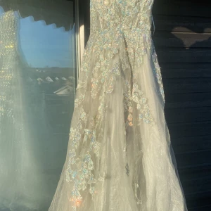 Ljusgrön långklänning med blommig spets och paljetter - Magisk ljusgrön långklänning med v-ringning och vackra blommiga spetsdetaljer i grönt och silver. Klänningen har glittrande paljetter och ett skirt, svepande tyg som ger en drömmig känsla. Perfekt för bal eller festliga tillfällen. Använd en gång, skriv om du har frågor kan gå ner i pris vid snabb affär