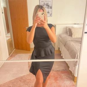 Svart klänning med volang och slits - Säljer en svart klänning med volangdetalj i midjan och slits framtill. Klänningen har korta ärmar och en snygg öppen rygg. Perfekt för dig som vill ha en stilren och elegant look.