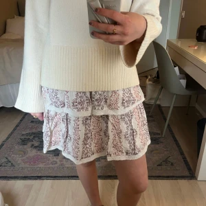 Vit och rosa mönstrad kjol med volang - Supersöt kortkjol i vitt med rosa blommönster. Kjolen har flera volanger och spetsdetaljer längs kanterna. Perfekt för en somrig look och lätt att matcha med en enkel topp.💞