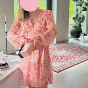 Rosa blommig kortklänning med volang - Superfin rosa kortklänning med blommigt mönster och volangdetaljer. Klänningen har långa ärmar med lätt puff och v-ringad hals. Perfekt för vår och sommar, och ger en romantisk känsla. Använd endast en gång 2024.
