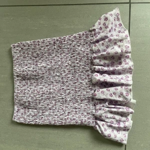 Vit och lila blommig kjol med volang - Säljer en söt kjol i vitt med lila blommönster och volangdetalj. Kjole är smockad för en tight passform och har en romantisk känsla. Perfekt för sommardagar!