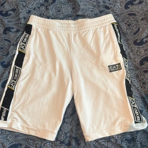 Vita shorts från Emporio Armani EA7 med svarta och gula detaljer - Snygga vita shorts från Emporio Armani EA7 med svarta och gula detaljer längs sidorna och logga på vänster ben. Elastisk midja med gul- och svartmönstrad snörning. Perfekta för en sportig och trendig look.