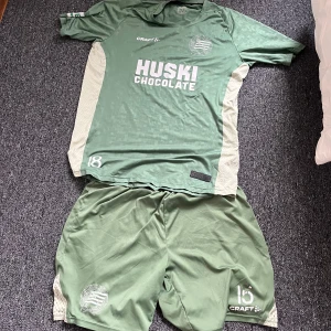 Grön fotbollströja från Craft Hammarby - Säljer en grön Hammarby fotbollströja från Craft med vita sidopaneler och tryck 'Huski Chocolate' på bröstet. Kortärmad modell med klubbmärke och Craft-logga. Perfekt för match eller träning.