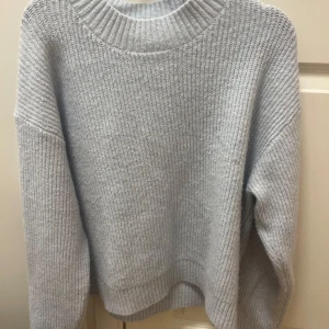 Ljusgrå stickad tröja från H&M - Mysig ljusblå stickad tröja från H&M med rund halsringning och ribbade detaljer vid hals, ärmslut och nederkant. Tröjan har en avslappnad passform och långa ärmar, perfekt för lager-på-lager under kyliga dagar. inte kommit till andvänding alls många gånger 